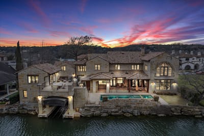 Twilight aerial view of the lakefront stone mansion with infinity pool, spa, and covered boat dock on Lake Austin
